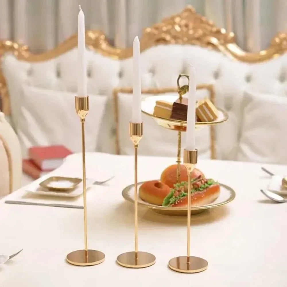A set of three gold-toned metal taper candle holders with white candles displayed on a white table.