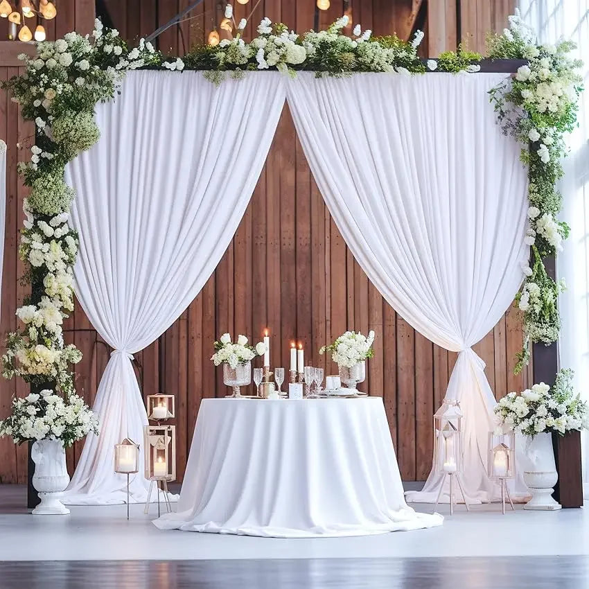 A white draped curtain backdrop decorated with greenery and white flowers for a wedding or event setting.