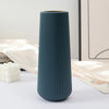 A tall, ribbed, dark teal plastic vase with a matte finish sitting on a white surface next to light beige fabric.