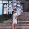A woman in a silver sequined dress stands on stone steps surrounded by clear and pink bubble balloons.
