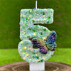 A large number five candle decorated with green sequins and a purple butterfly on a white base.