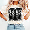 A woman wearing a light beige t-shirt with a black silhouette design featuring witches and a moon.