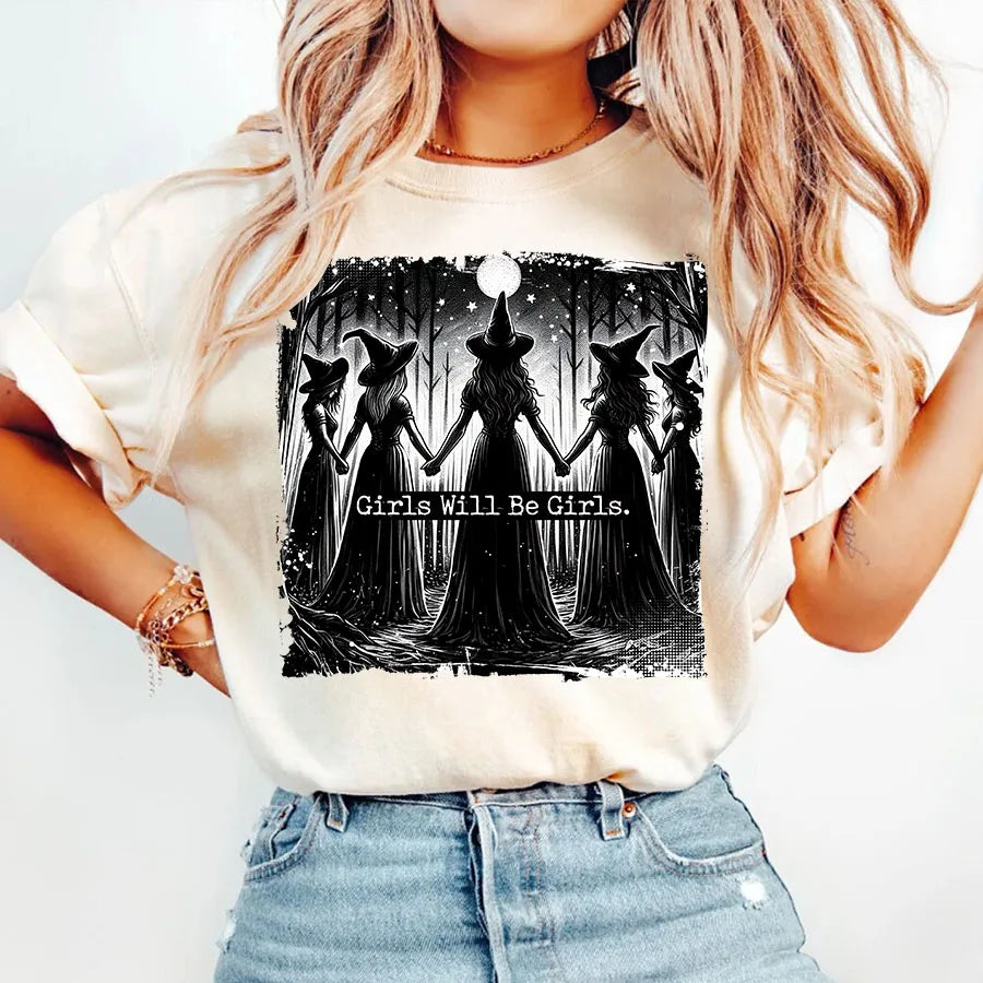 A woman wearing a light beige t-shirt with a black silhouette design featuring witches and a moon.