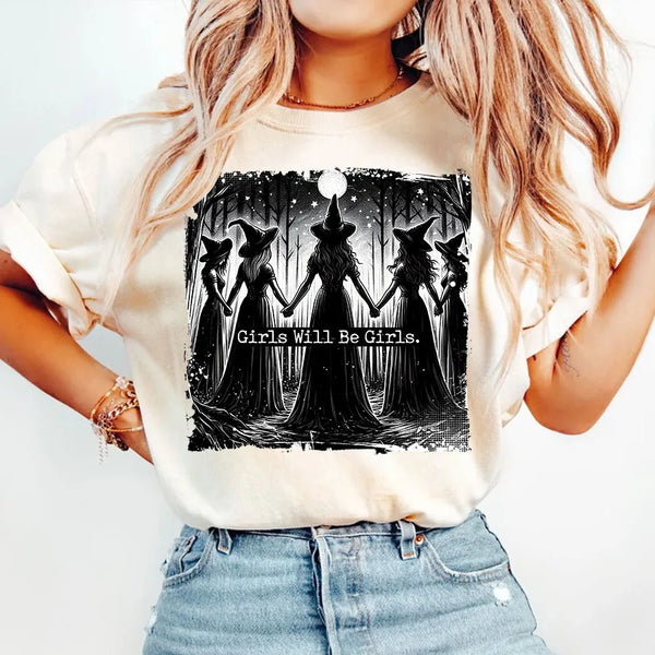 A woman wearing a light beige t-shirt with a black silhouette design featuring witches and a moon.
