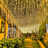 Una densa cortina de luces blancas cálidas cuelga sobre un camino de jardín y vegetación.