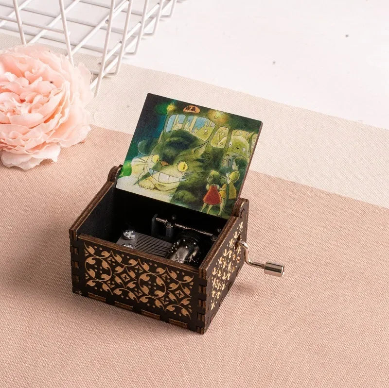 A hand-cranked wooden music box featuring a Totoro design, resting on a light pink surface.