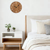 A minimalist wooden wall clock hangs on a white wall above a light wood nightstand next to a bed.