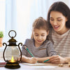 A woman is sitting with a young girl, both writing at a desk near a vintage battery-powered desk lamp.