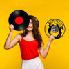 A smiling woman holds a black vinyl record and a vintage wall clock with a silhouette of a tiger on its face against a yellow