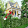 A bright yellow house with a porch and white railing is framed by a dense green artificial ivy hedge fence.
