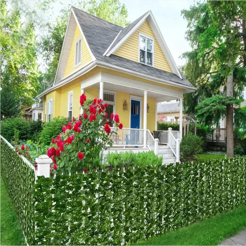 A bright yellow house with a porch and white railing is framed by a dense green artificial ivy hedge fence.