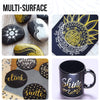A collection of multi-surface art featuring black and gold paint pens on various surfaces like stones, paper, and a mug.