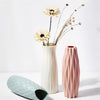 A white vase holds dried flowers and delicate stems against a bright background, next to a pink textured vase.