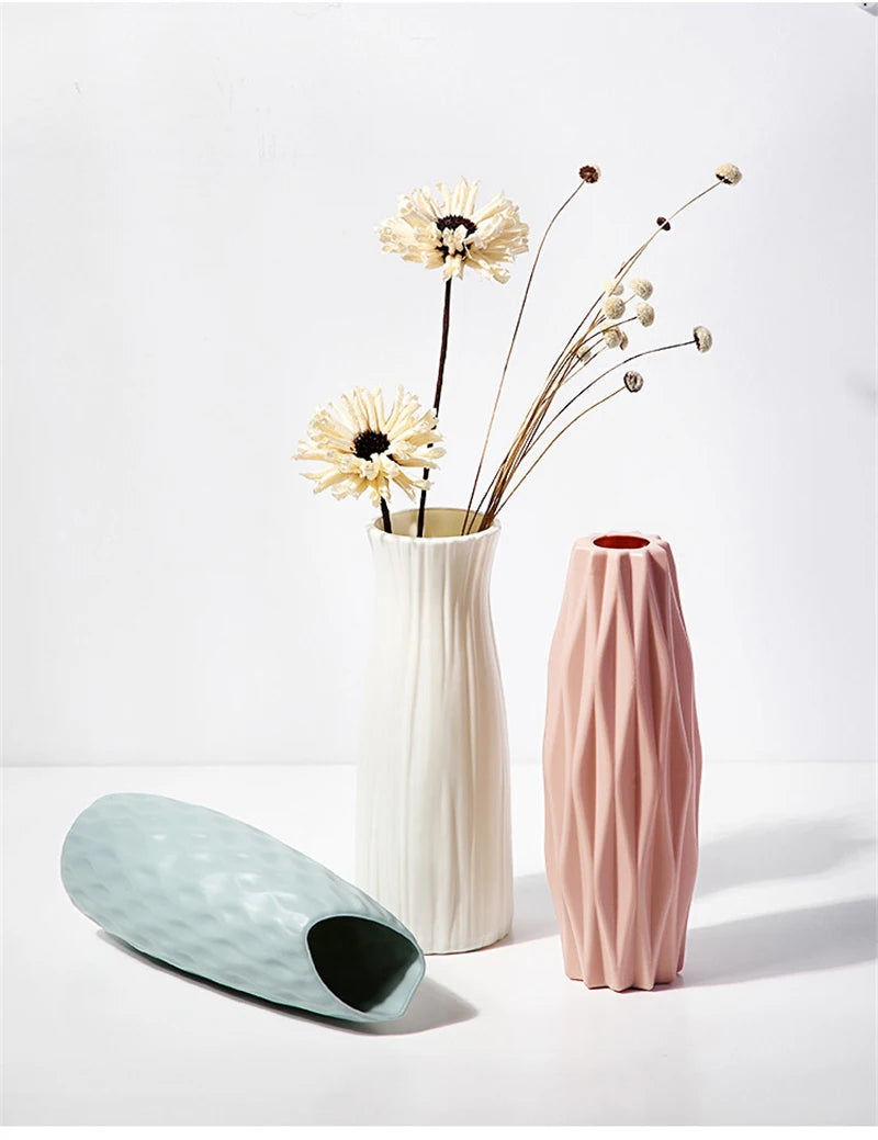 A white vase holds dried flowers and delicate stems against a bright background, next to a pink textured vase.