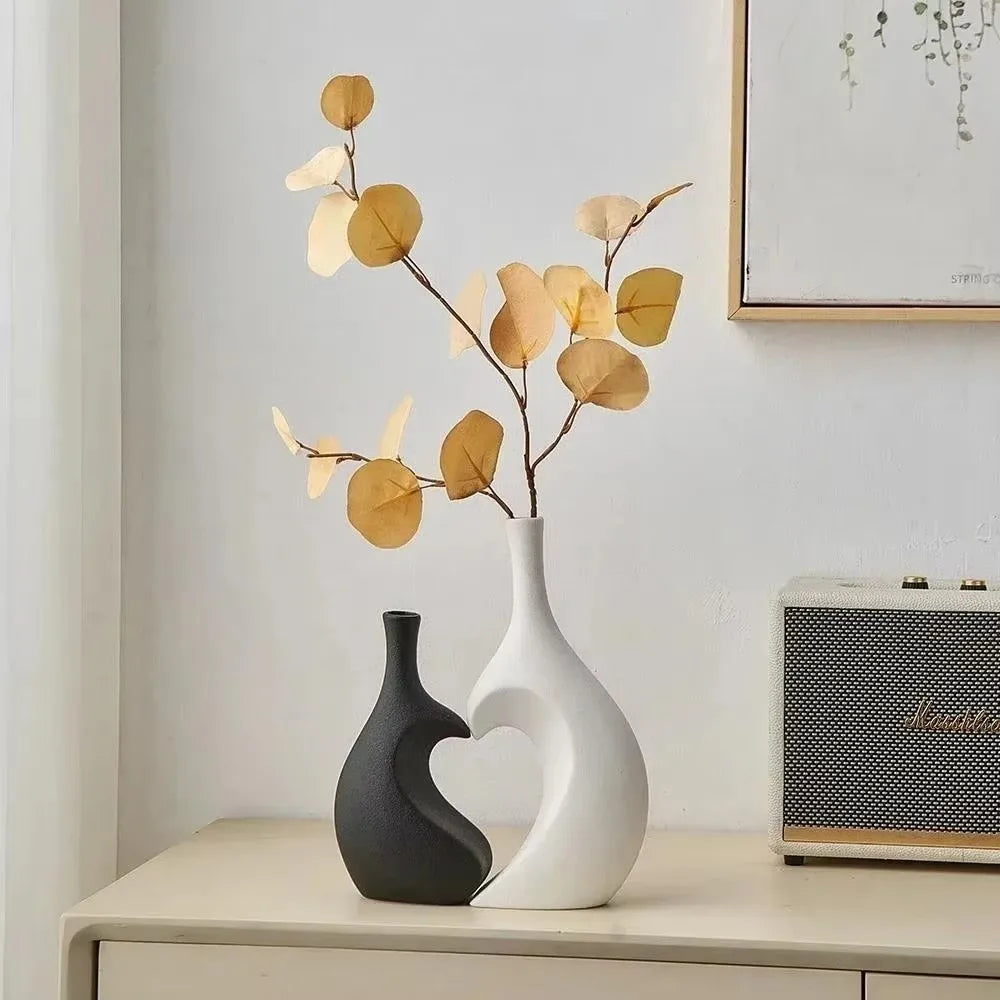 A modern heart-shaped ceramic vase holds delicate dried leaves against a white wall.