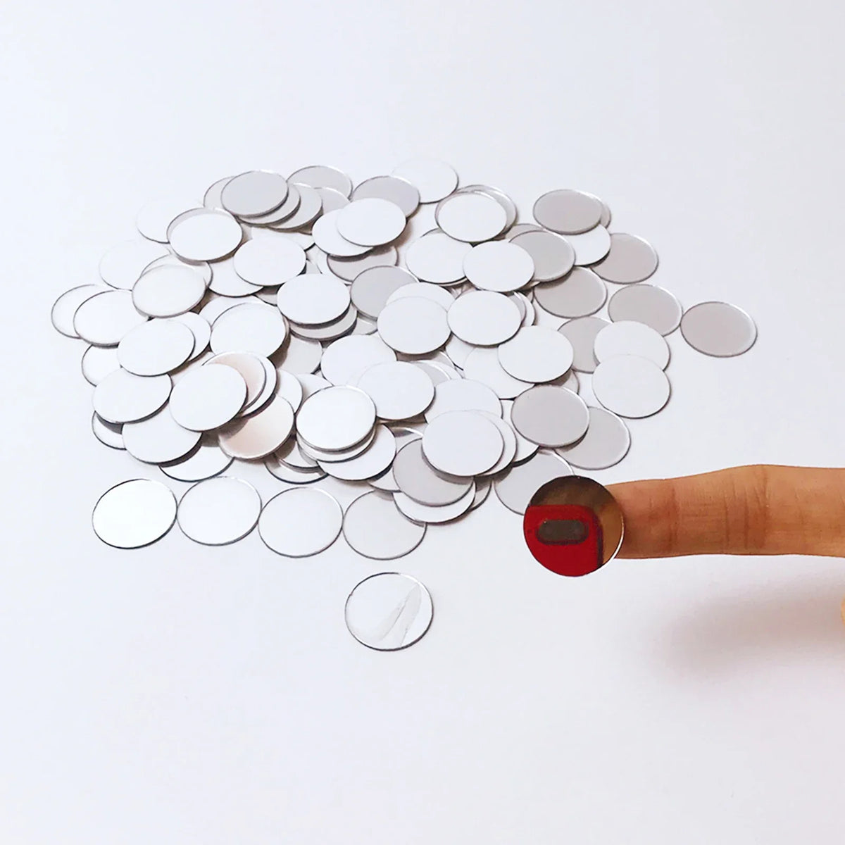 A hand is holding a pile of round silver mirror tiles against a white background.