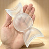 A hand holds a clear natural selenite triple moon charging bowl against a light wooden background.