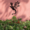 Black metal wall art featuring a silhouette of a dragon against a pink wall with green foliage in the foreground