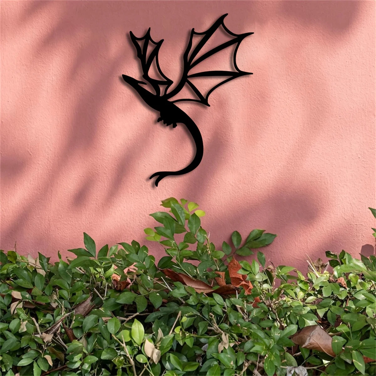Black metal wall art featuring a silhouette of a dragon against a pink wall with green foliage in the foreground