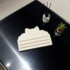 A white cat face squeezer tool is placed on a black tray next to a small potted plant and a black stick.