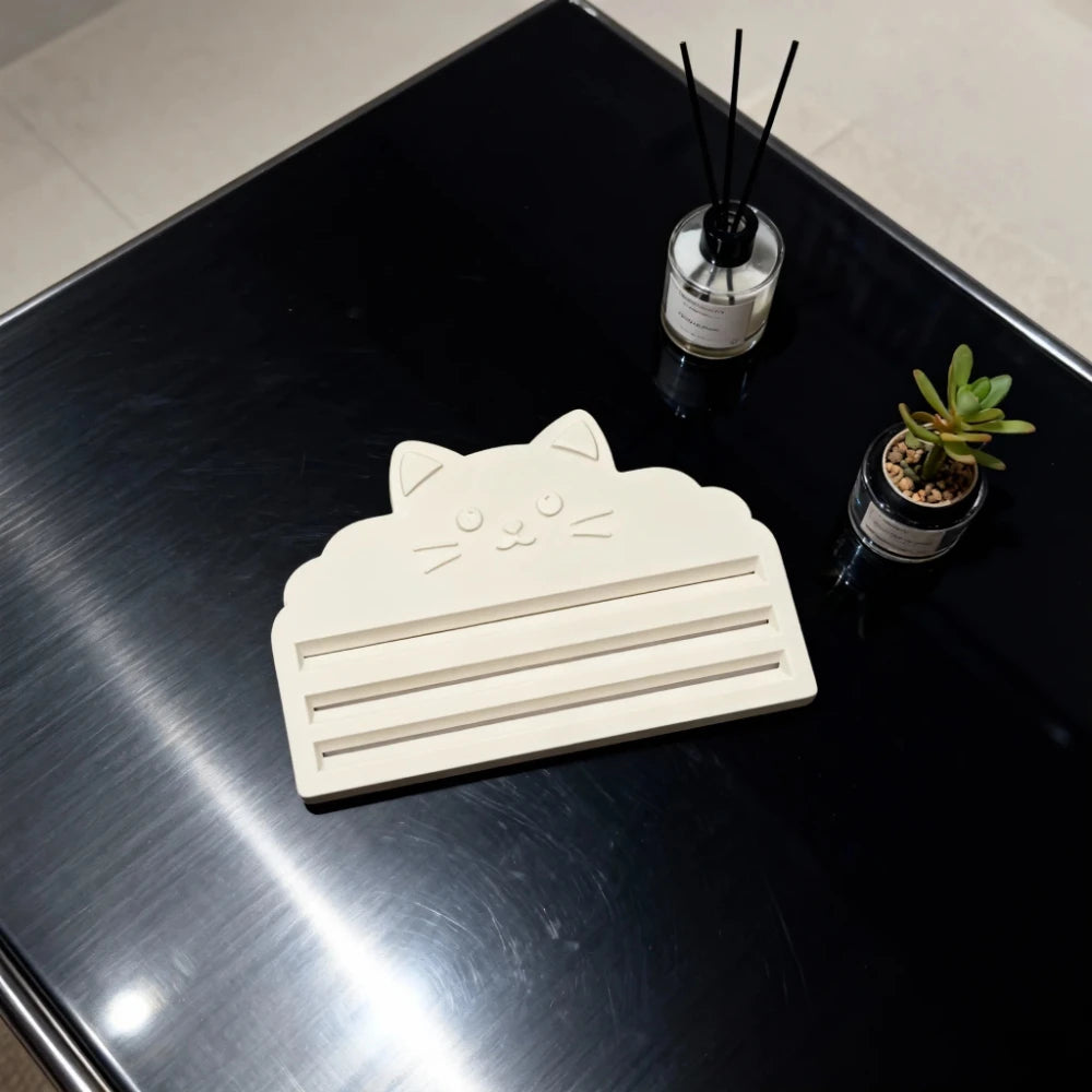 A white cat face squeezer tool is placed on a black tray next to a small potted plant and a black stick.