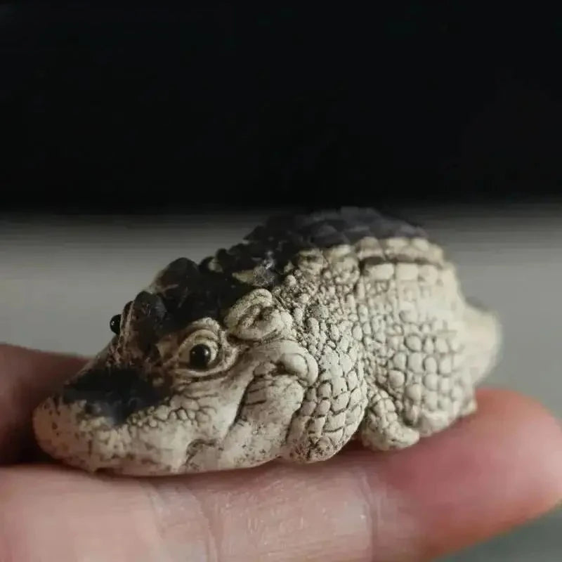 A small, textured resin crocodile tea pet ornament is held in a hand against a dark background.