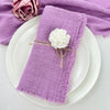 A purple fringed cotton napkin is placed on a white plate, with a white flower decoration and a purple cloth in the backgroun