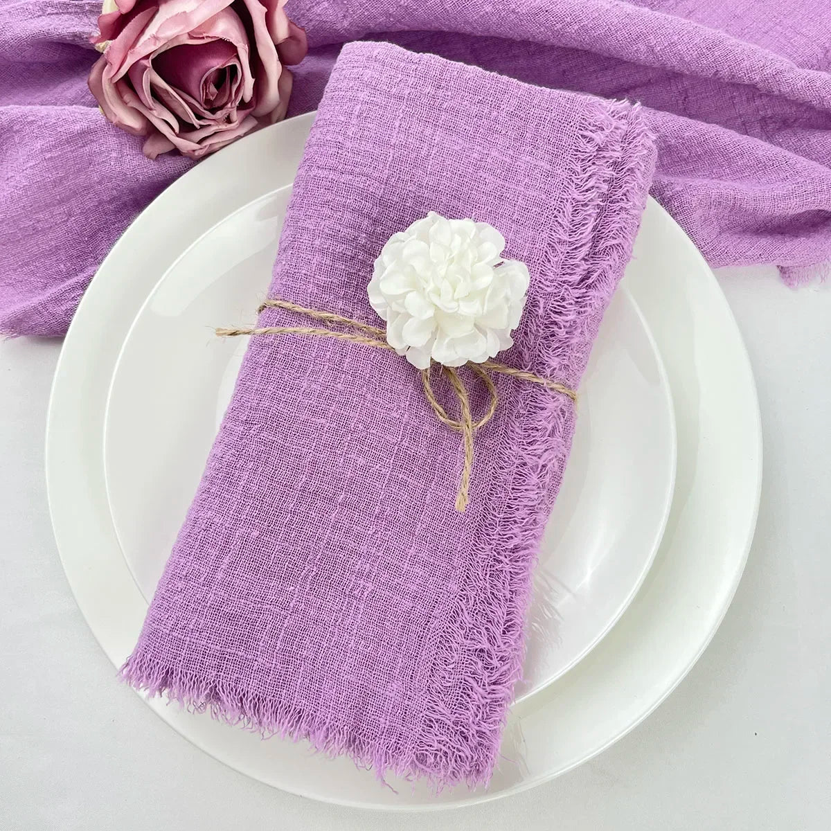 A purple fringed cotton napkin is placed on a white plate, with a white flower decoration and a purple cloth in the backgroun