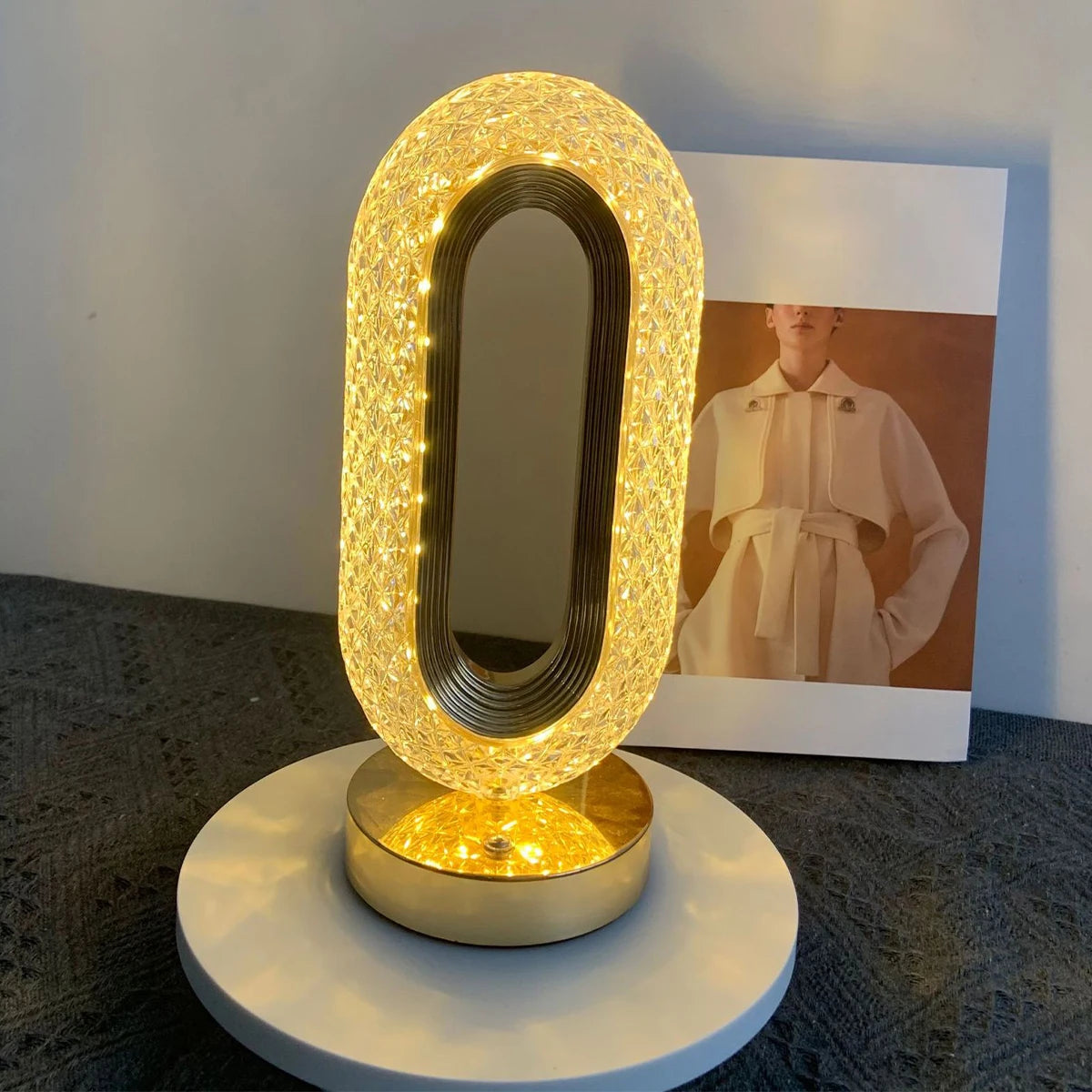 An oval crystal table lamp with a glowing light and mirror is displayed on a white base.