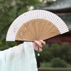 A person holds an elegant bamboo folding fan with a tassel, displaying the fan against a blurred outdoor background.