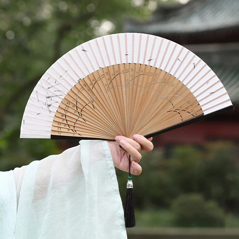 A person holds an elegant bamboo folding fan with a tassel, displaying the fan against a blurred outdoor background.