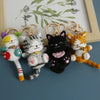 Four handcrafted crochet cat keychain pendants in various colors and textures displayed on a wooden frame.