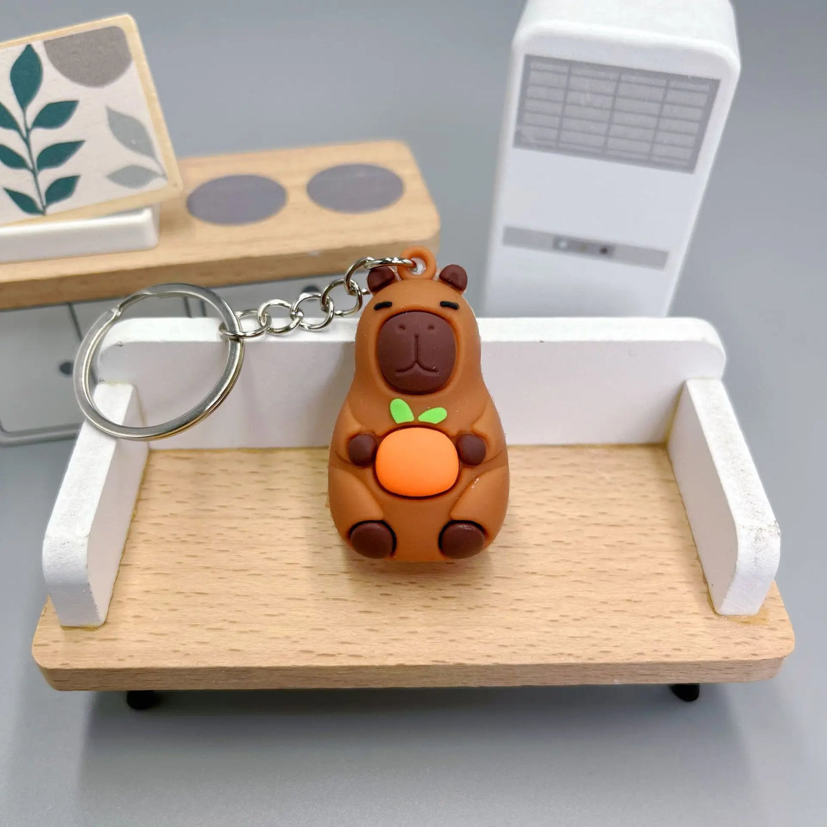 Un petit pendentif de clé en forme de capybara brun rond posé sur un plateau en bois clair avec une boîte rectangulaire blanc