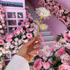 A hand holds a delicate, gold-stemmed artificial crystal flower against a backdrop of pink roses and white decor.
