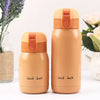 Two beige stainless steel vacuum insulated coffee mugs with orange lids are displayed next to some greenery.