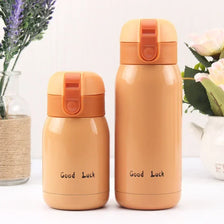 Two beige stainless steel vacuum insulated coffee mugs with orange lids are displayed next to some greenery.