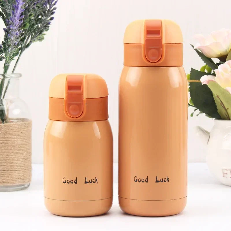 Two beige stainless steel vacuum insulated coffee mugs with orange lids are displayed next to some greenery.