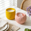 A yellow candle jar and a pink textured silicone mold sit on a white surface next to a white napkin and silverware.
