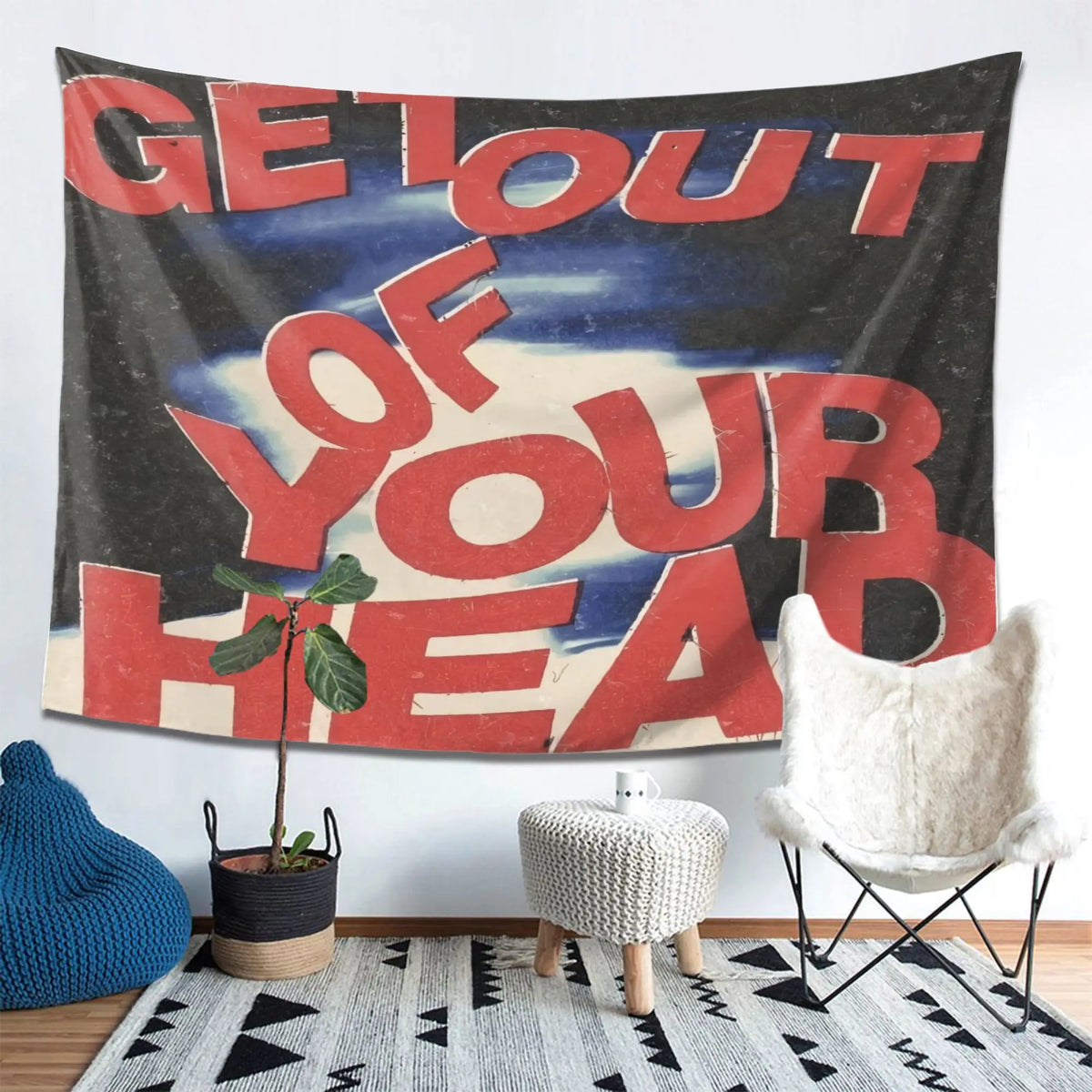 A dark tapestry wall hanging with red text that reads Get Out of Your Head displayed above a rug and a white chair.