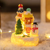 A glowing Christmas gingerbread house resin ornament set featuring a snowman and a decorated gingerbread structure in snow.