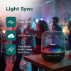 A transparent RGB Bluetooth speaker is displayed on a wooden table in a dimly lit room with people in the background.
