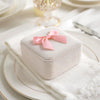 A square white box with a pink satin ribbon bow resting on a white napkin on a plate.