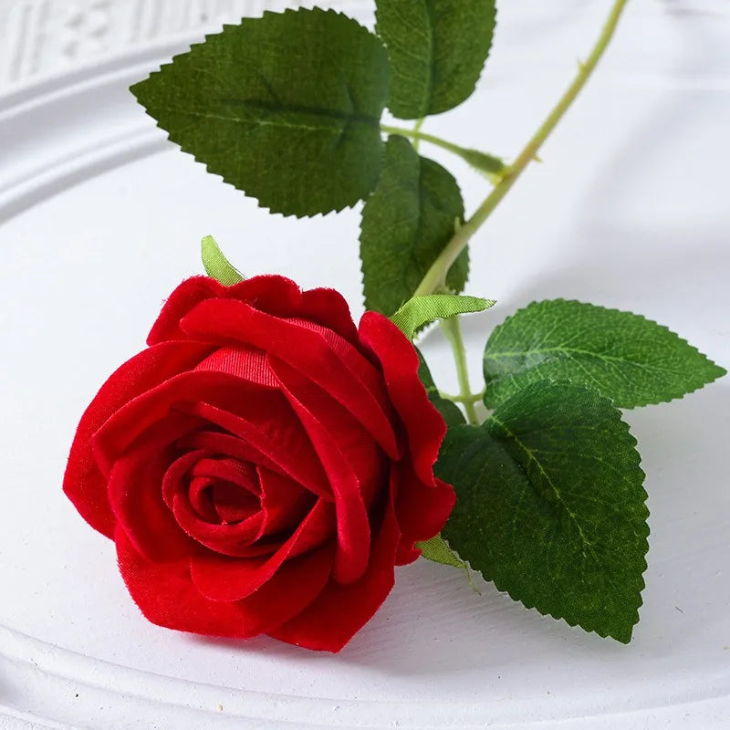 A vibrant red velvet artificial rose with green leaves on a white background.