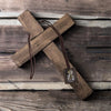 A wooden cross pendant necklace set with a leather cord and a small tag on a rustic wooden surface.
