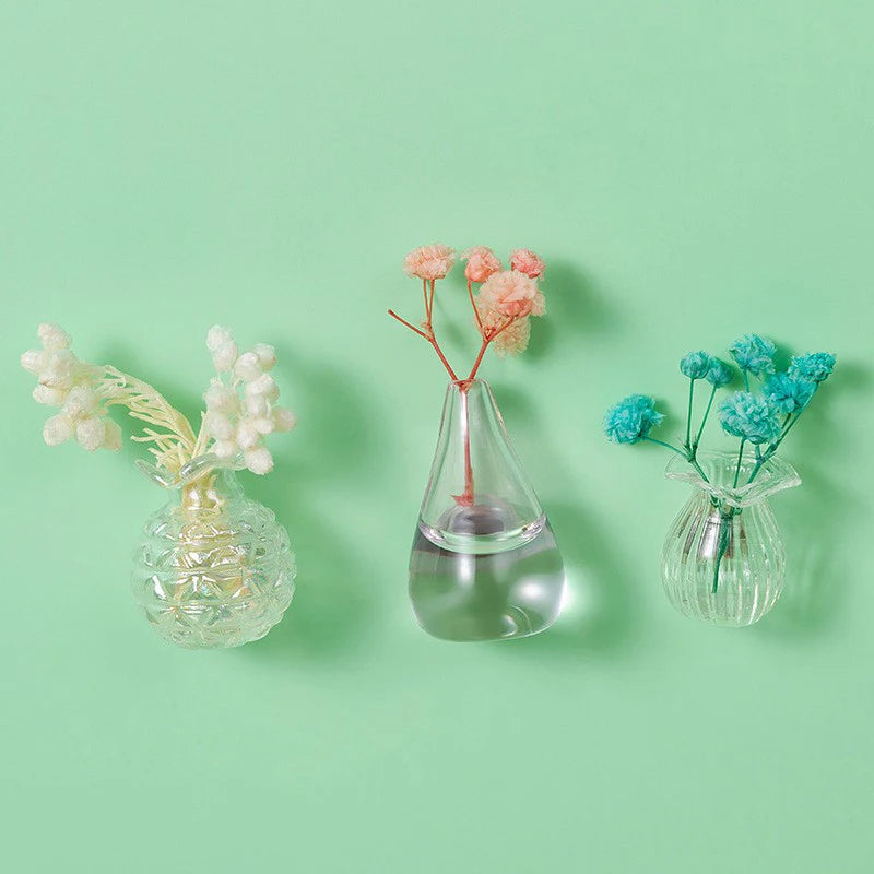Three glass vials hold dried pink and blue flowers against a light green background.