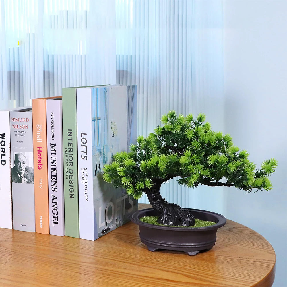 A green artificial bonsai tree in a dark pot sits on a wooden table next to several books.