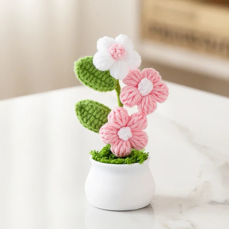 Hand woven crochet flowers in pink and white are potted in a white ceramic container.