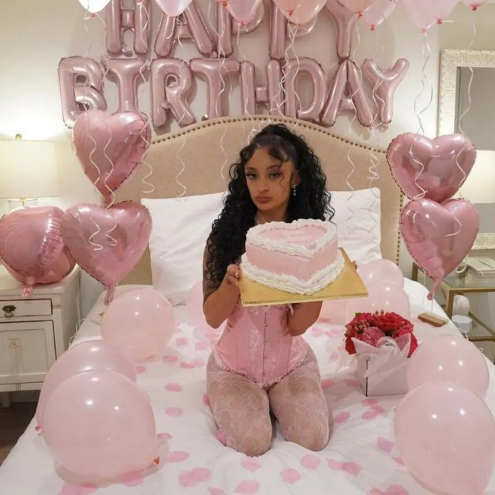 A woman in pink attire is sitting on a bed decorated with pink balloons and a cake, surrounded by foil letter balloons.