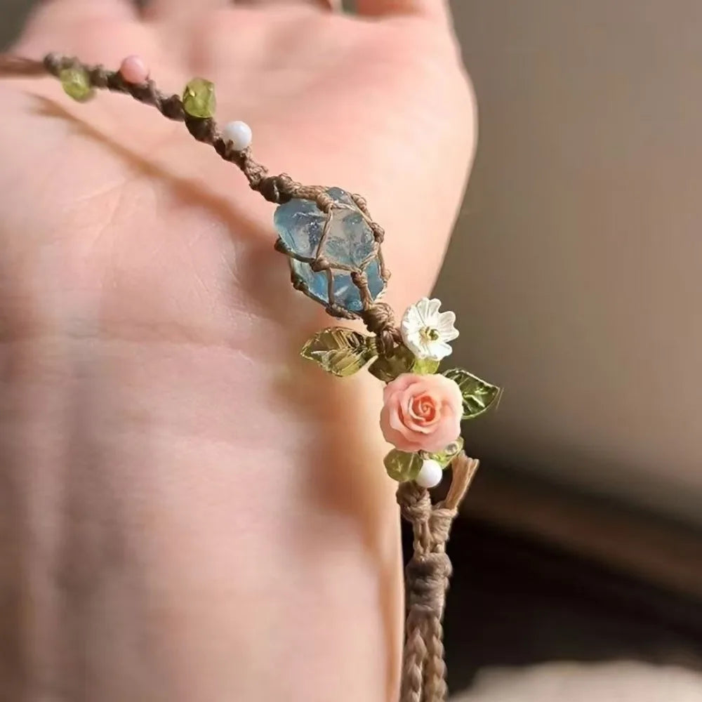 A hand holds a macrame bracelet featuring raw aquamarine beads, small white pearls, and a pink rose accent.
