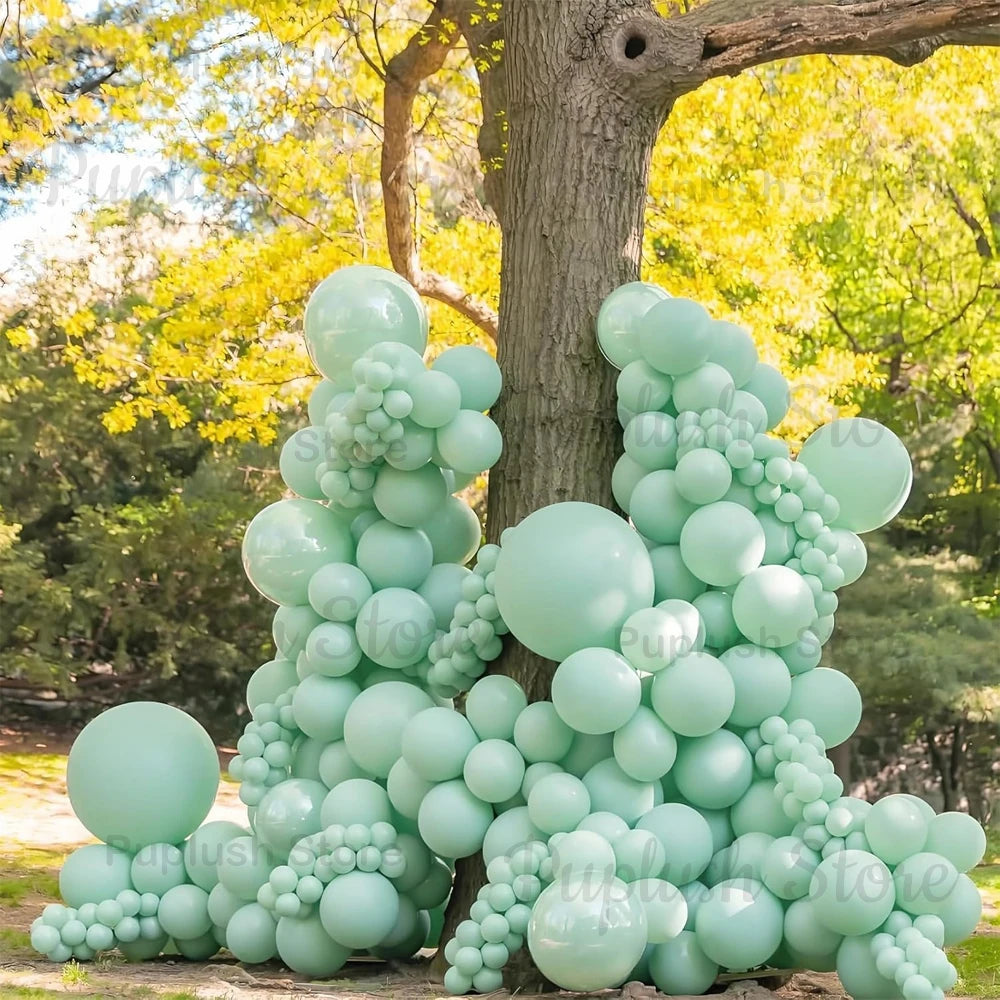 A large arrangement of sage green latex balloons clustered around a tree trunk in an outdoor setting.